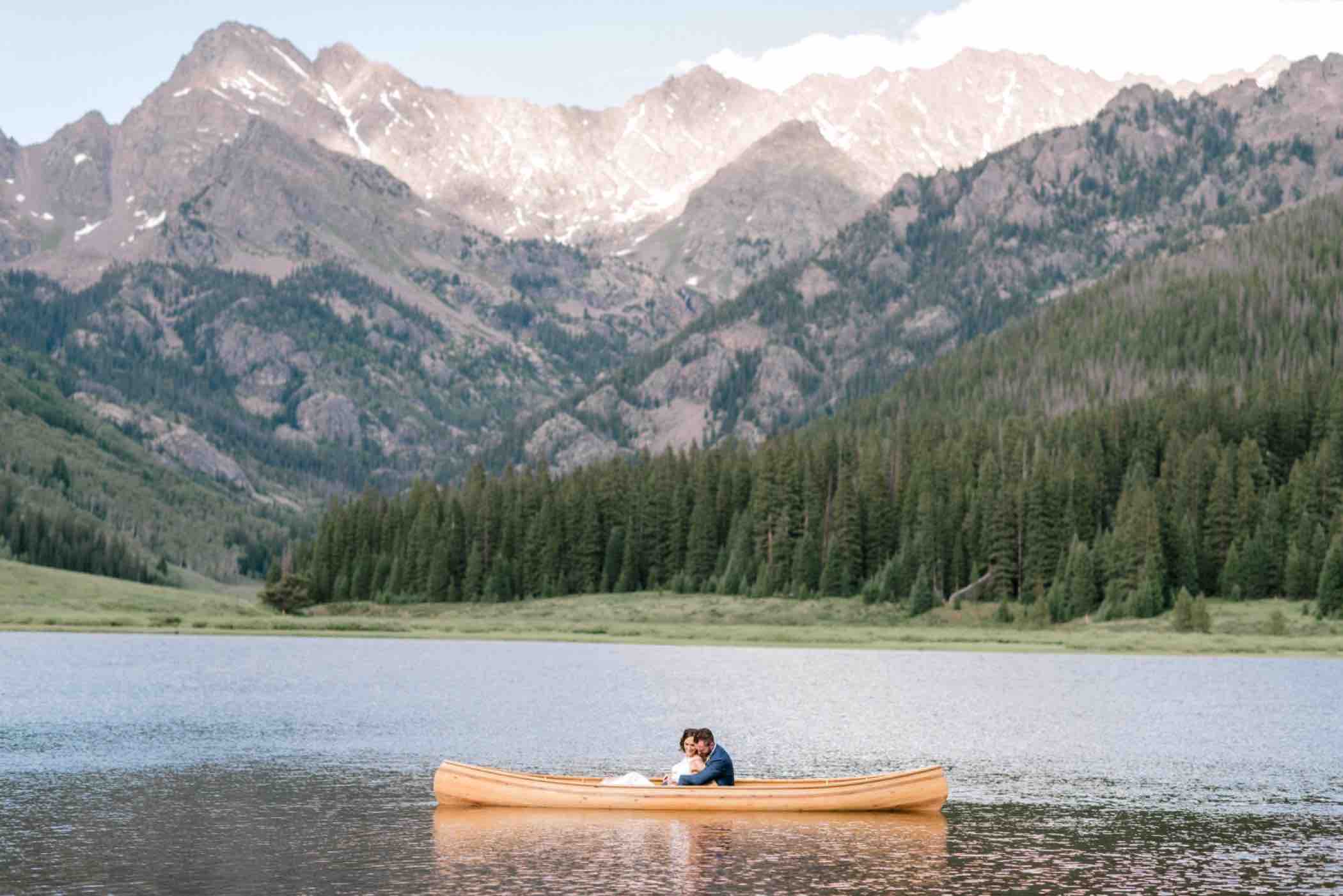 The Rocky Mountains are visible from Piney Lake at Piney River Ranch. Bride and groom photos in a wooden canoe in Vail Colorado. Photo by Ali and Garrett, Romantic, Adventurous, Nostalgic Wedding Photographers.