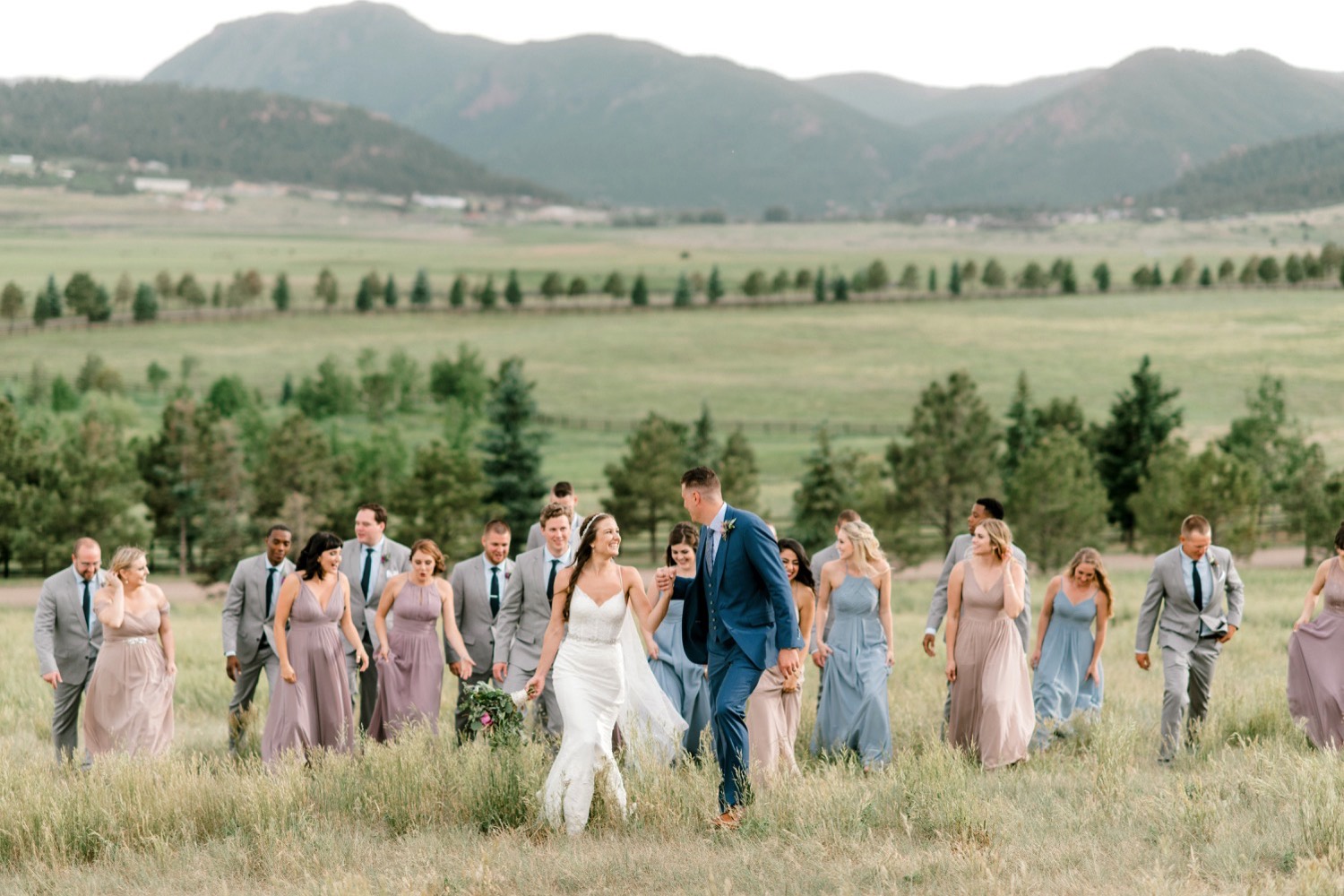 Kristen and Charlton are joined by their wedding party with the groomsmen in their blue ties and the bridesmaids in their purple, blush, and blue gowns as they walk through the field of Spruce Mountain Ranch in Larkspur, Colorado.