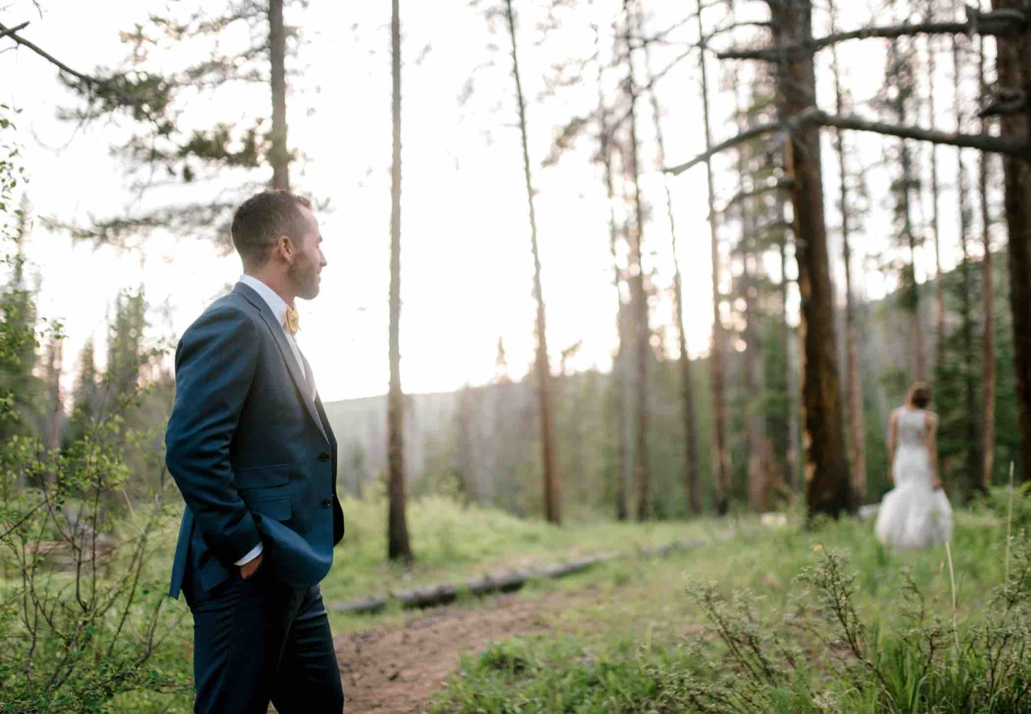 Kris the groom watches his bride Sallie walk through the pine forest outside Piney River Ranch in Vail, Colorado. Photo by Ali and Garrett, Romantic, Adventurous, Nostalgic Wedding Photographers.